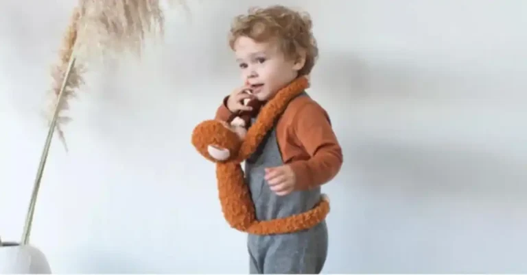 A toddler wearing a plush crochet spider monkey as a scarf, standing next to a decorative plant.